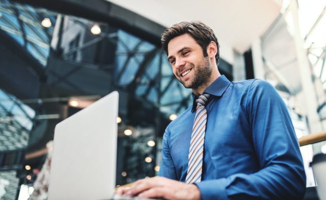 a-young-businessman-sitting-on-a-bench-in-a-modern-building-using-laptop--e1635759348322